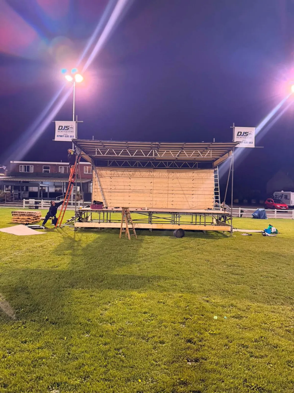 Two scaffolders building a covered scaffold stage with a timber back wall on a floodlit grass field at night, DJS banners on both corners