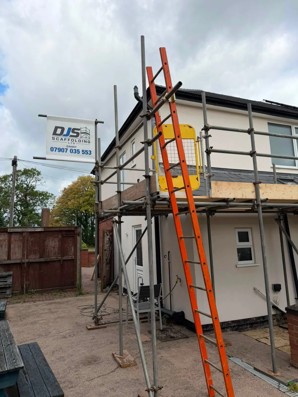 Single-lift DJS scaffold at first-floor level across the back of a cream rendered house, with an orange ladder, yellow safety gate and DJS banner